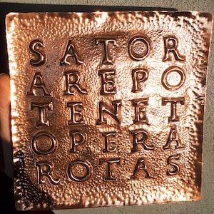 May include: A square, copper-colored plaque with the Latin word square palindrome "SATOR AREPO TENET OPERA ROTAS" embossed in capital letters. The plaque has a textured surface and is held by a person.