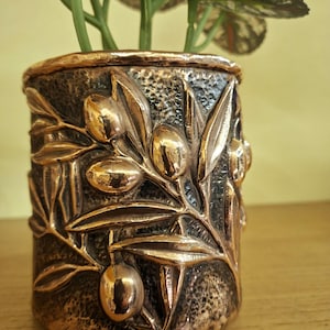 May include: A bronze-coloured, cylindrical planter with an embossed olive branch design. The planter has a textured background and a shiny, metallic finish on the olives and leaves. Green plant stems and leaves emerge from the top.