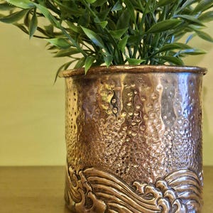 May include: A copper-colored metal planter with a textured surface and a wave design at the base. The planter holds green leafy plants. The planter has a cylindrical shape and sits on a wooden surface.