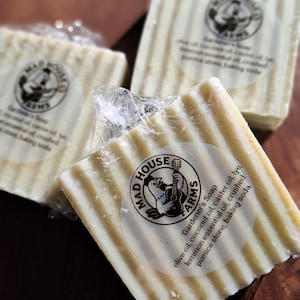May include: Close-up of three bars of Gardener's Soap from Mad House Farms. The bars are wrapped in clear plastic and have a light yellow color with vertical ridges. Each bar has a circular label with a rooster playing a guitar.