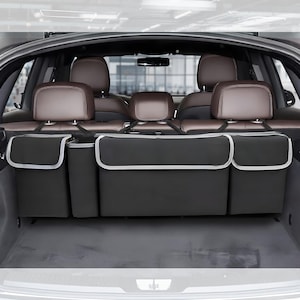 May include: A black car trunk organizer with three compartments and a silver trim. The organizer hangs from the headrests of the back seat. The car's interior features brown leather seats.