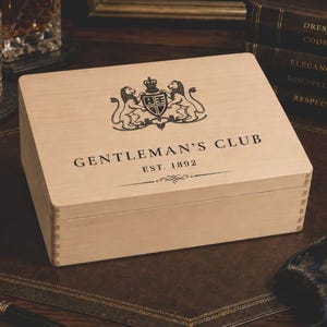 Könnte beinhalten: Eine rechteckige, helle Holzschatulle mit Klappdeckel, verziert mit einem schwarzen Wappen aus zwei Löwen, die einen Schild flankieren, sowie dem Text GENTLEMAN'S CLUB und EST. 1892. Die Schatulle hat an den Ecken sichtbare Fingerzinken.