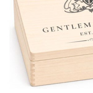 Peut inclure: Une bo&icirc;te en bois clair avec des coins &agrave; joints dent&eacute;s. Le couvercle pr&eacute;sente un graphique imprim&eacute; en noir repr&eacute;sentant un blason au lion et le texte partiel GENTLEM EST. La bo&icirc;te a une finition en grain de bois naturel et une forme rectangulaire.