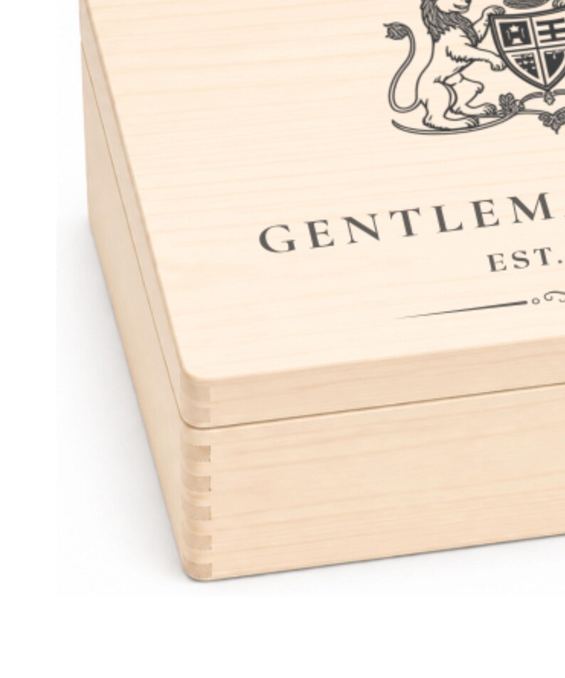 Peut inclure: Une bo&icirc;te en bois clair avec des coins &agrave; joints dent&eacute;s. Le couvercle pr&eacute;sente un graphique imprim&eacute; en noir repr&eacute;sentant un blason au lion et le texte partiel GENTLEM EST. La bo&icirc;te a une finition en grain de bois naturel et une forme rectangulaire.