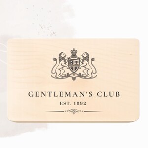 Peut inclure: Une bo&icirc;te rectangulaire en bois clair pr&eacute;sentant un motif imprim&eacute; marron fonc&eacute; compos&eacute; de deux lions entourant un blason central surmont&eacute; d'une couronne. Sous l'embl&egrave;me, le texte indique GENTLEMAN'S CLUB, EST. 1892, avec une ligne d&eacute;corative.