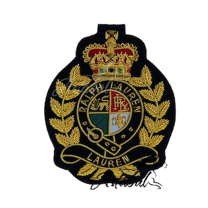 Pode incluir: Um patch bordado preto com detalhes dourados e vermelhos. O patch apresenta uma coroa, folhas de louro e as palavras "RALPH LAUREN" em um design circular. O centro tem um escudo com um design multicolorido.