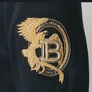 May include: Close-up of black fabric with a gold embroidered patch. The patch features a stylized eagle, the letter "B", and the word "BALMAIN" in a circular design. The word "PARIS" is also visible.