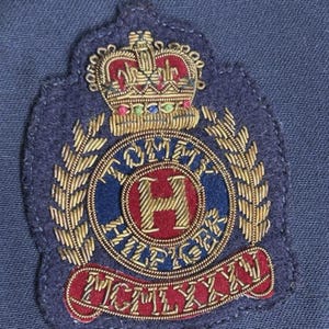 May include: A navy blue embroidered patch featuring a gold crown and laurel leaves. The patch has the words "TOMMY HILFIGER" and "MCMLXXXI" in gold lettering. The center features a red "H" within a blue circle.