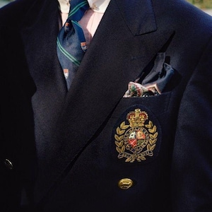 May include: A navy blue blazer with a crest patch, gold buttons, and a patterned pocket square. A striped tie and collared shirt are visible. The blazer has a classic, tailored design.