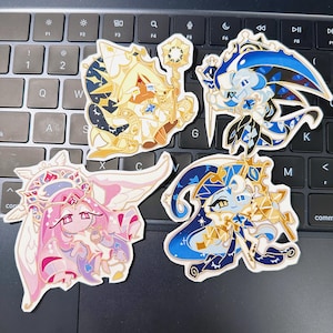 May include: Four colorful character stickers on a laptop keyboard. The stickers feature cartoon characters in gold, blue, and pink with intricate designs. One character holds a staff. The stickers are likely for fans of a specific game or series.