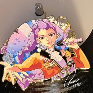 May include: A colorful enamel pin featuring a stylized illustration of a person with purple hair holding a microphone. The pin has a black jacket, orange and red clothing, and the text "Rrrrmm". The pin is set against a black background.