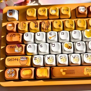 May include: A custom keyboard with yellow and white keycaps. The keycaps feature honey-themed designs, including bees, honeycombs, and jars of honey. The keyboard has a yellow frame and the text "ORGANIC HONEY" on the spacebar.