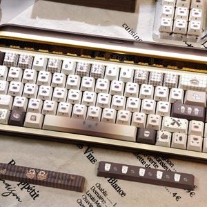 May include: A custom mechanical keyboard with white and brown keycaps. The keycaps feature bear faces and coffee cup designs. Additional keycaps and a wrist rest are also visible.