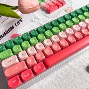 May include: A gradient-colored mechanical keyboard with green, pink, and red keycaps. The keyboard has a clear plastic frame and a red base. The keys are arranged in a standard layout, with the text "OURISM LIFE" visible in the background.