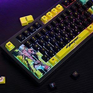 Puede incluir: Teclado mecánico negro con teclas amarillas y negras. El teclado presenta la palabra "Cyberpunk" en la barra espaciadora y un gráfico colorido en las teclas de función. Las teclas están retroiluminadas con un arcoíris de colores.