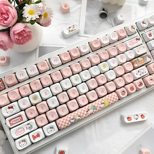May include: A white mechanical keyboard featuring custom keycaps with a pastel pink and white color scheme. The keycaps are decorated with various Hello Kitty character graphics, including bows, cupcakes, and stars, with a checkered spacebar.