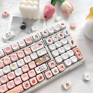 Pode incluir: Um teclado mecânico branco com teclas rosa e brancas. As teclas apresentam várias ilustrações, incluindo personagens Hello Kitty, flores e alimentos. O teclado tem um layout padrão com teclas de função e um teclado numérico.