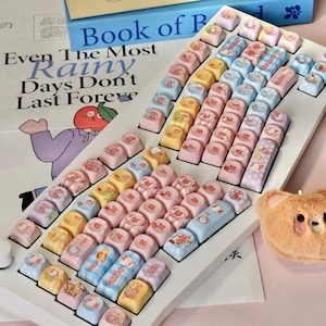 May include: A custom keyboard with pastel-colored keycaps. The keycaps feature various designs, including cartoon characters, patterns, and text. The keyboard is on a white base, with a plush bear and a book in the background.
