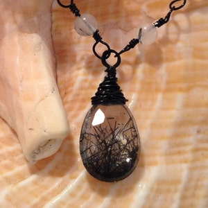 May include: A black wire wrapped necklace with clear quartz beads and a black tourmaline pendant. The necklace is displayed on a seashell.