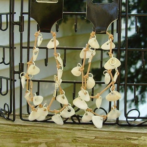 Op de afbeelding: Twee kettingen gemaakt van bruin lederen koord en witte schelp hangers. De kettingen hangen aan een zwarte metalen display.