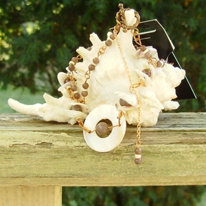 Könnte beinhalten: Eine goldene Kettenkette mit braunen Perlen und einem großen weißen Muschelanhänger. Die Kette wird auf einer Muschel präsentiert.