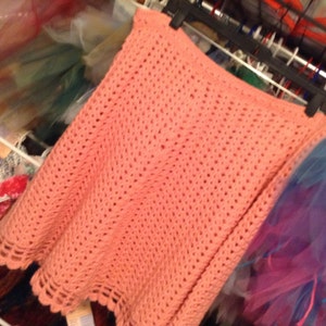 Pode incluir: Uma saia de crochê de cor pêssego claro com uma bainha festonada. A saia é feita com um padrão de ponto simples e tem um caimento solto e fluido.