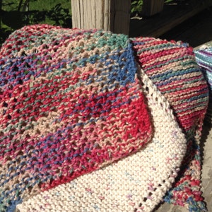 May include: A stack of colorful knitted dishcloths in shades of pink, blue, green, and beige. The cloths are made with a variety of stitches and textures.