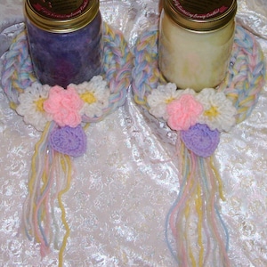 May include: Two crocheted candle rings with pastel pink, yellow, and white flowers and purple accents. The rings are made with a braided yarn and have long, colorful yarn tails.
