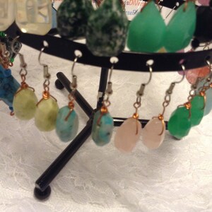 Puede incluir: Una muestra de pendientes con diversas cuentas de piedras preciosas y semipreciosas. Los pendientes están en un soporte negro con un gancho plateado y un alambre de cobre.