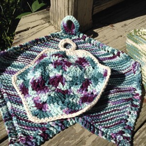 Puede incluir: Dos paños de cocina de punto en tonos de morado, turquesa y blanco. Un paño de cocina es cuadrado con un borde de ganchillo y un asa de bucle. El otro paño de cocina es rectangular con un patrón de rayas.