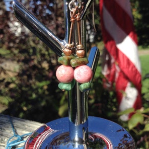 Op de afbeelding: Een paar oorbellen met roze en groene edelsteen kralen. De oorbellen hangen aan een zilveren standaard.