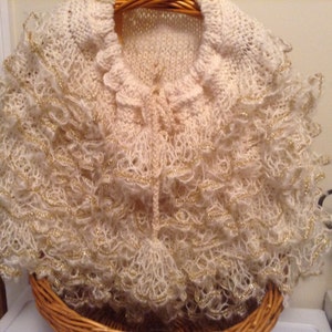 May include: Cream-colored knitted shawl with ruffled edges and gold metallic thread accents. The shawl features a delicate, open-weave design and a tie closure at the front. The garment is displayed in a woven basket.