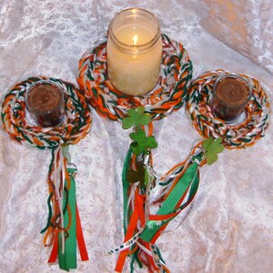 May include: Three white, green, and orange braided rope rings with a white ribbon tied to each. The rings are decorated with shamrocks and hold small votive candles.