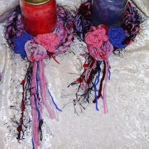 May include: Two crocheted candle rings with colorful yarn tassels. The rings are decorated with pink and blue crocheted flowers.