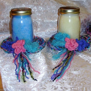 May include: Two glass jars with lids, one filled with blue wax and the other with white wax, are displayed on crocheted yarn bases with colorful yarn tassels.