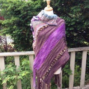 Puede incluir: Un poncho de punto a rayas moradas y marrones con flecos. El poncho tiene un cuello alto y está decorado con adornos marrones.