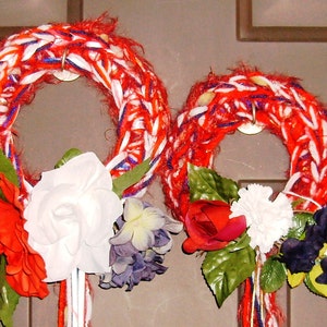 May include: Two red, white, and blue yarn wreaths with white, red, and blue ribbons hanging from the bottom. The wreaths are decorated with artificial flowers, including white roses, red roses, white carnations, and blue flowers.