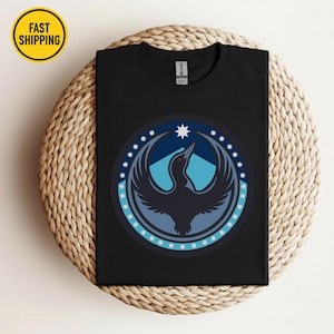 May include: Black t-shirt featuring a circular design with a bird-like figure in shades of blue and a white star. The design is surrounded by a blue border with white stars. The shirt is displayed on a woven surface.