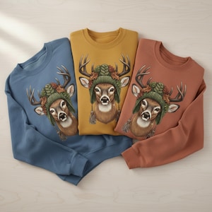 Pode incluir: Três camisolas de gola redonda nas cores azul, amarelo mostarda e ferrugem. Cada camisola apresenta um gráfico de veado usando um gorro de malha verde com folhas de outono. O veado tem grandes chifres e um rosto detalhado.