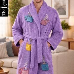 Op de afbeelding: Een lila badjas van badstof met geborduurde koffiekopjes. De badjas heeft een sjaalkraag, een tailleband en zakken. De tekst "The LeatherOxide" staat in de linkerbovenhoek.