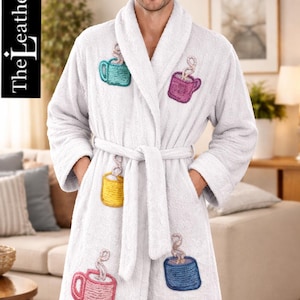 Puede incluir: Bata de rizo blanca con diseños de tazas de café bordadas en varios colores. La bata tiene cuello chal, cinturón y bolsillos. El texto "The Leather Oxide" está impreso verticalmente en el lado izquierdo de la imagen.