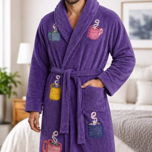 May include: A plush, purple bathrobe with embroidered coffee cup designs in various colors. The robe has a shawl collar, a waist tie, and pockets. The embroidered cups are blue, pink, yellow, and blue. The robe is made of a soft, absorbent material.