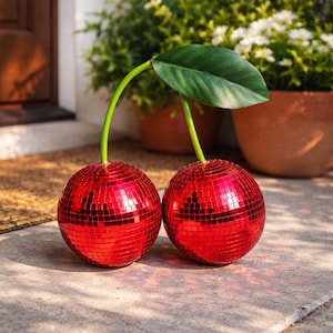 May include: Two red disco ball cherries with green stems and a single green leaf. The cherries are made of small, reflective squares. The decorative fruit is placed on a concrete surface, with potted plants in the background.
