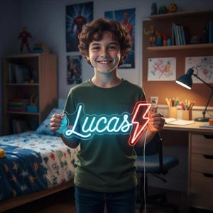 Puede incluir: Una persona sonriente sostiene un letrero de neón con el nombre "Lucas" en letras azules y un rayo rojo. La persona lleva una camiseta verde. La habitación tiene una cama, un escritorio y carteles.