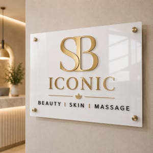 May include: A rectangular white acrylic sign with gold metallic lettering reading SB ICONIC and black text reading BEAUTY | SKIN | MASSAGE. The sign features a small gold leaf icon and is mounted with four circular gold standoffs at the corners.