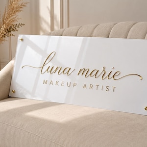 Könnte beinhalten: Ein rechteckiges Schild aus weißem Acryl mit goldfarbenen Schriftzügen, die "luna marie" in Schreibschrift und "MAKEUP ARTIST" in einer serifenlosen Schrift zeigen. Das Schild ist mit vier goldfarbenen, runden Abstandshaltern befestigt.