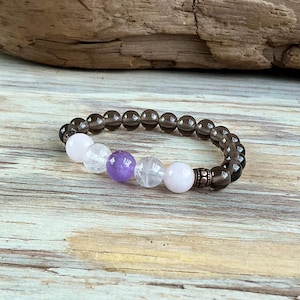 May include: A beaded bracelet featuring a series of translucent dark brown smoky quartz beads, interspersed with a central arrangement of two pale pink rose quartz beads, two clear quartz beads, and one vibrant purple amethyst bead.