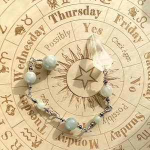 May include: A handcrafted bracelet with pale blue gemstone beads and a clear crystal pendant. The bracelet is strung on silver-coloured wire, displayed on a wooden divination board with astrological symbols and text.
