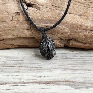 May include: A black, irregularly shaped stone pendant encased in a dark, woven cord macrame cage. The pendant hangs from a matching black cord necklace, featuring a textured, glossy surface with subtle grey inclusions throughout the stone.