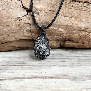 May include: A necklace featuring a teardrop-shaped stone with a mottled black and silver metallic surface. The stone is secured in a black macrame cord netting that connects to a thin black cord, creating a simple pendant design.